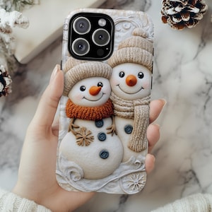 May include: A phone case featuring a 3D design of two snowmen. The snowmen wear knitted hats and scarves in shades of beige and orange. The case has a white, textured background with decorative patterns and is held by a person.