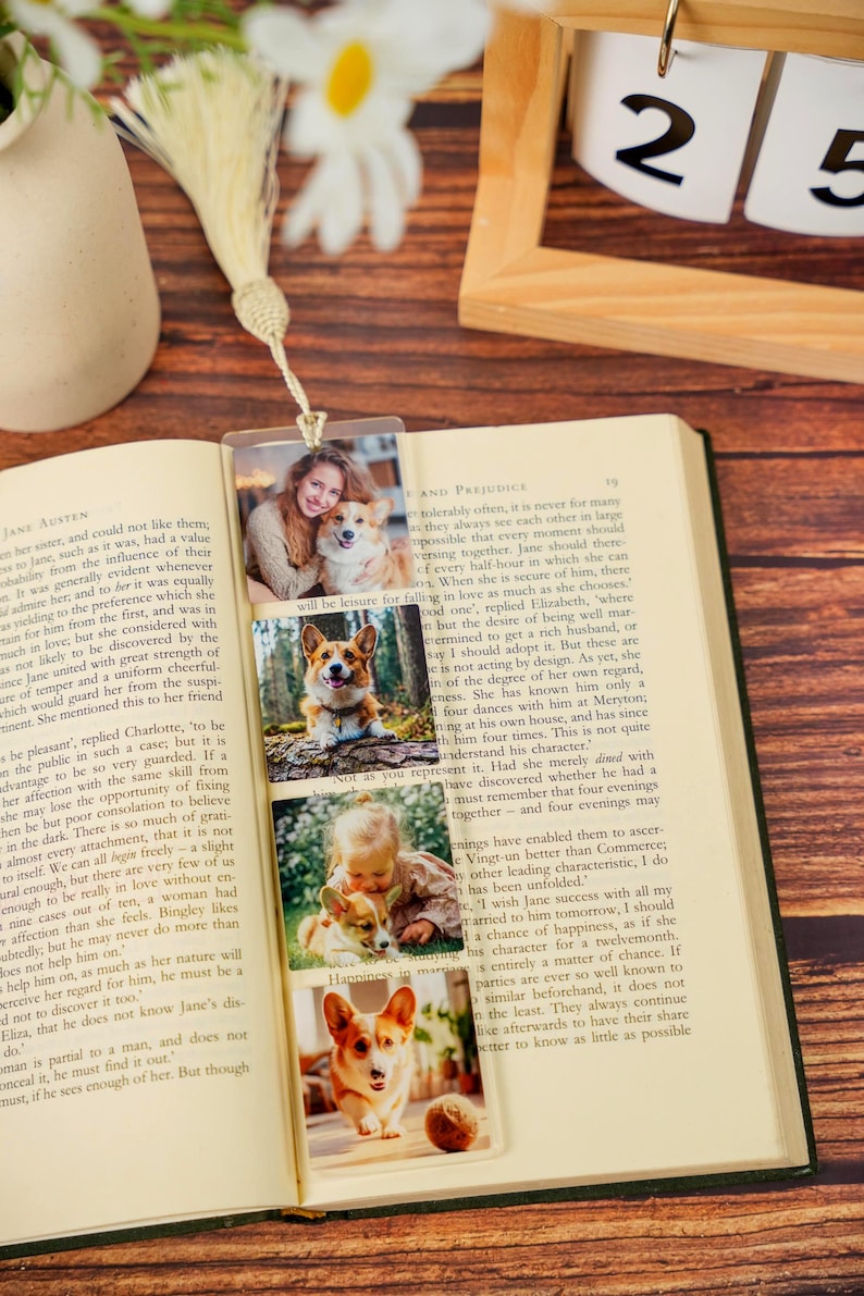 May include: A clear rectangular bookmark featuring four stacked photographic prints of a person and a corgi dog. The bookmark is topped with a cream-colored fabric tassel and is placed inside an open book with printed text on the pages.