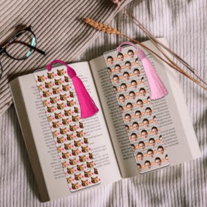 May include: Two clear bookmarks with pink tassels rest on an open book. One bookmark features a pattern of cat faces and hearts, while the other displays a pattern of faces and flowers. The book is open on a neutral-toned surface.