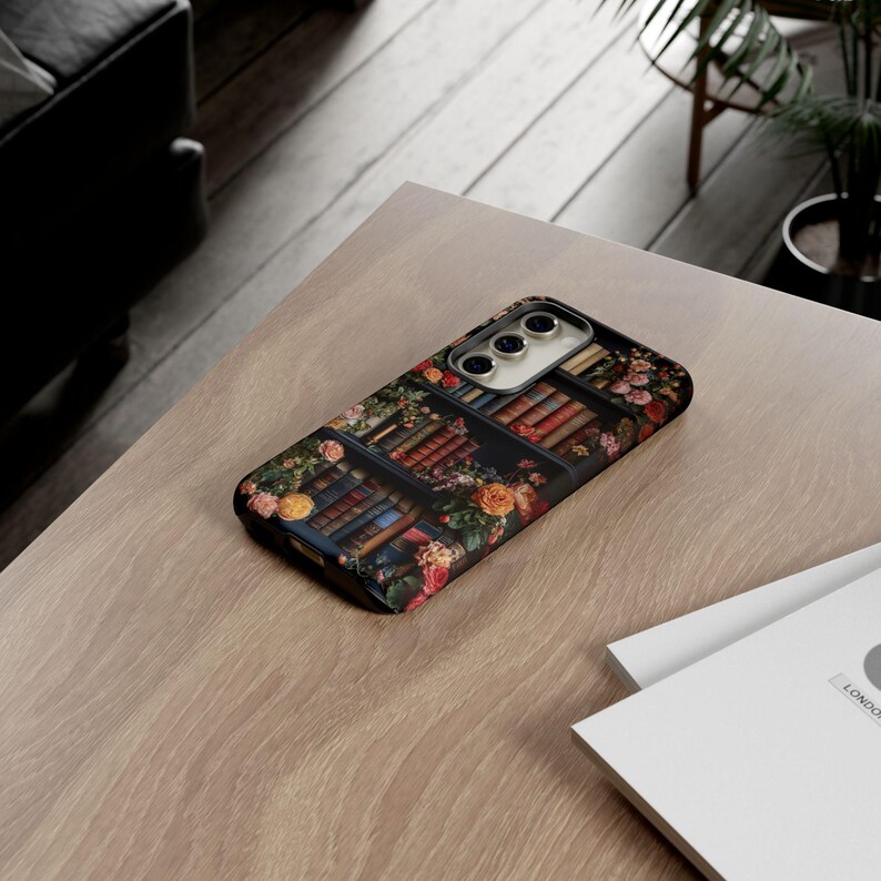 Puede incluir: Una funda de tel&eacute;fono negra con un dise&ntilde;o floral que presenta una estanter&iacute;a llena de libros. La funda tiene un recorte para el objetivo de la c&aacute;mara y un borde elevado alrededor de los bordes.