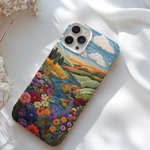 May include: A phone case with a colorful embroidered design of a landscape with a river, hills, and flowers. The case has a white background and a camera lens cutout.