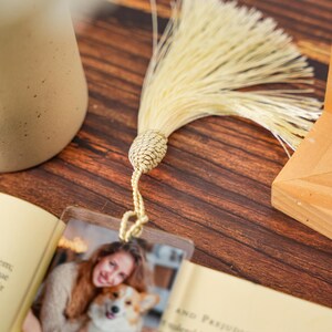 May include: A rectangular clear acrylic bookmark featuring a printed photograph of a person holding a dog. The bookmark is attached to a long, cream-colored decorative tassel with a braided knot at the top, resting on an open book page.