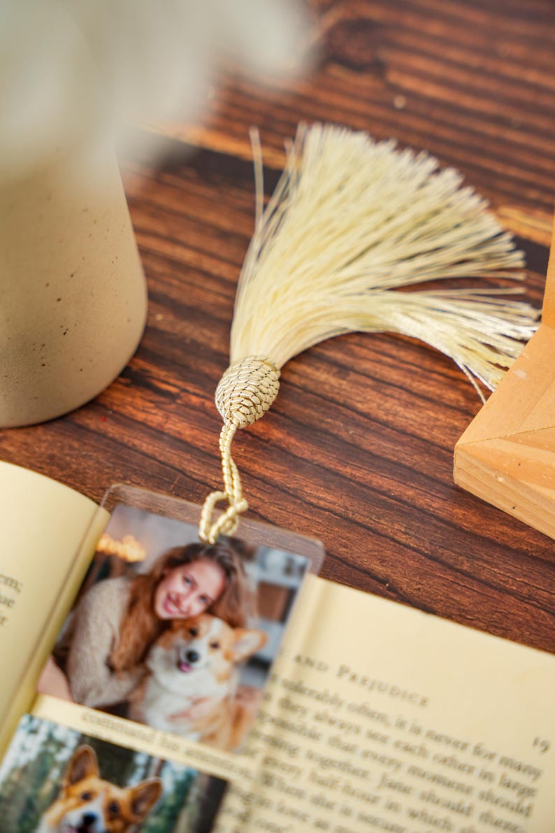 May include: A rectangular clear acrylic bookmark featuring a printed photograph of a person holding a dog. The bookmark is attached to a long, cream-colored decorative tassel with a braided knot at the top, resting on an open book page.