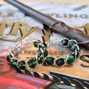 Peut inclure: Boucles d'oreilles créoles argentées ornées de perles vert foncé, une baguette et un sablier doré sont présentés sur un livre. Le livre a des lettres dorées et un fond rouge. La baguette est marron.