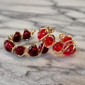 Peut inclure: Deux bagues faites à la main avec des perles rouges et du fil d'or. Les perles sont d'un rouge profond et le fil est d'un or brillant. Les bagues sont posées sur une surface en marbre.