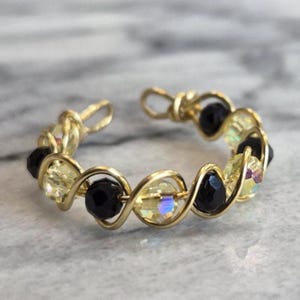 May include: An open-style ring crafted from gold-toned wire, featuring a repeating pattern of alternating faceted black beads and iridescent, light-reflecting clear beads. The wire is twisted into loops that secure each bead in a continuous circular band.