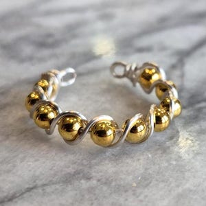 May include: A ring featuring a silver-colored wire base wrapped in a spiral pattern with small, polished gold-colored spherical beads. The band is open at the top, showcasing a continuous row of metallic beads secured by the coiled wire structure.