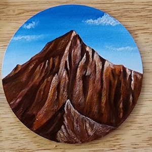 Puede incluir: Un posavasos redondo pintado a mano con un paisaje de montaña. La obra representa una cordillera marrón bajo un cielo azul con nubes blancas. El posavasos es de madera y tiene un acabado brillante.