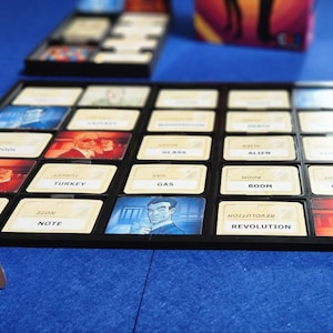 May include: A black game board with 16 squares, each containing a card with a word or image. The words include "Note", "Turkey", "Gas", "Boom", "Revolution", and "Mercury". The images include a man in a suit, a woman with long hair, and a man with a mustache.