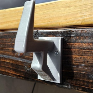 May include: A gray, 3D-printed hook mounted on a dark wooden surface. The hook has a rectangular base and a curved arm, secured with a single screw. The hook is designed to hold items and is attached to a wooden surface.