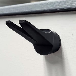 May include: A black plastic hook with two prongs, mounted on a white wall.