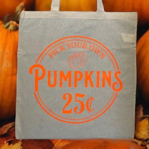 May include: A gray canvas tote bag with orange text that reads "Pick Your Own Pumpkins 25¢". The bag is surrounded by orange pumpkins.