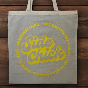 May include: A white tote bag with a yellow circle design that says "Side Chick" and lists various food items around the circle. The food items listed are: "Yams, Cranberry Sauce, Mashed Potatoes, Green Bean Casserole, Stuffing, Gravy".