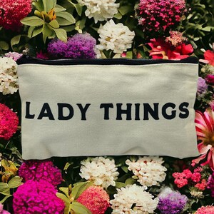 May include: A rectangular, off-white canvas cosmetic bag with the words "LADY THINGS" printed in black. The bag has a black zipper and is surrounded by a vibrant array of pink, red, purple, and white flowers.
