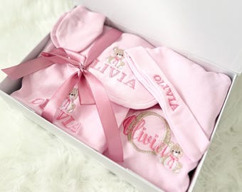 May include: A pink baby gift set in a white box, tied with a pink ribbon. The set includes a hat, bib, headband, and a onesie, all embroidered with the name "Olivia" and a teddy bear design.