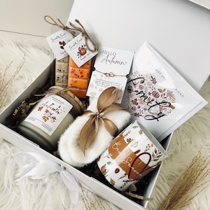 May include: A white gift box filled with autumn-themed items. Includes a candle, wax melts, a mug, a bracelet, and a sachet. The box is tied with a white ribbon and contains decorative elements like dried leaves and a fluffy white slipper.