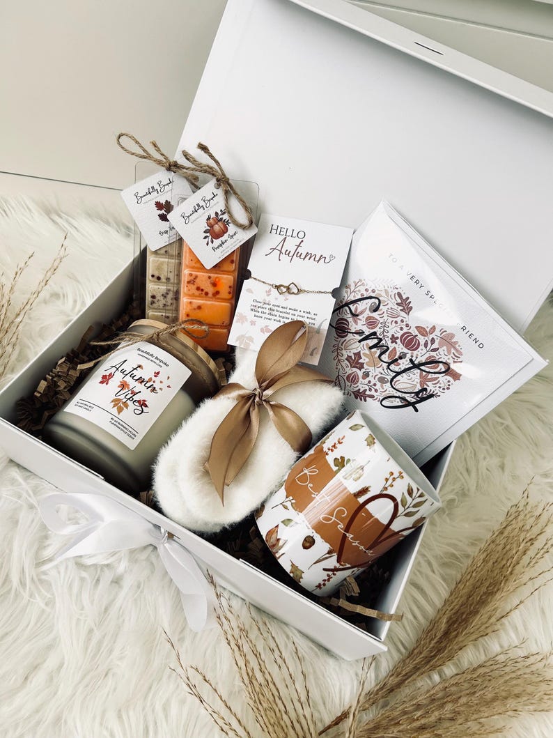 May include: A white gift box filled with autumn-themed items. Includes a candle, wax melts, a mug, a bracelet, and a sachet. The box is tied with a white ribbon and contains decorative elements like dried leaves and a fluffy white slipper.