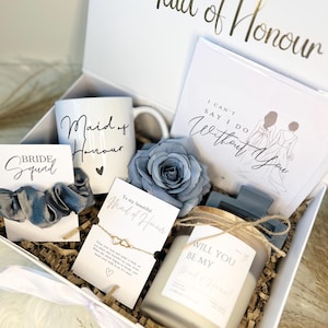 May include: A white gift box containing a white mug with Maid of Honour text, a blue fabric rose, a card with I Can't Say I Do Without You text, a grey scrunchie on a Bride Squad card, a gold-tone bracelet on a card, and a candle with a label.