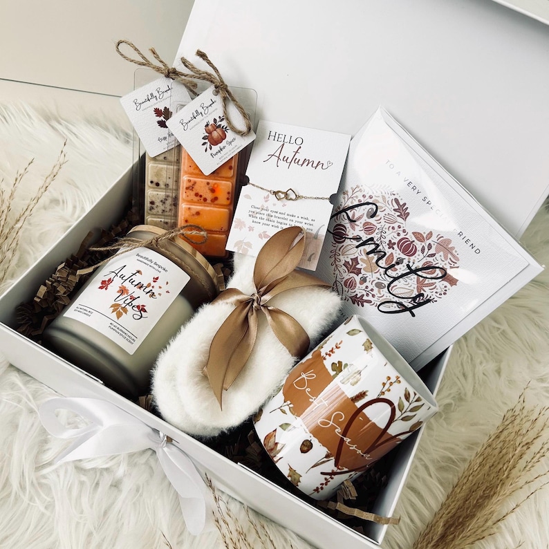 May include: A white gift box filled with autumn-themed items. Includes a candle, wax melts, a mug, a bracelet, a pair of slippers, and a card. The box is tied with a white ribbon and contains decorative paper.