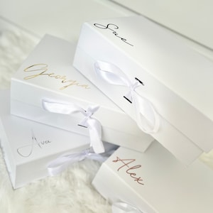 May include: Four white gift boxes with white satin ribbon bows. The boxes are stacked on top of each other. The top box has the name "Sue" written on it in black ink. The second box has the name "Georgia" written on it in gold ink. The third box has the name "Ava" written on it in black ink. The bottom box has the name "Alex" written on it in rose gold ink.
