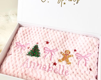May include: A white gift box with a pink blanket inside. The blanket has embroidered bows, a Christmas tree, a gingerbread man, and the name "Annabella". The box lid has "Merry Christmas" in gold script.