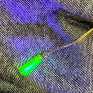 May include: A neon green pendant necklace with a gold-colored chain. The pendant is a smooth, elongated shape. The necklace is displayed on a textured, gray and black fabric.