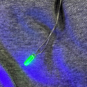 May include: A silver necklace with a glowing green cylindrical pendant. The pendant emits a bright green light against a textured gray and blue background. The chain is delicate and the pendant is the focal point.