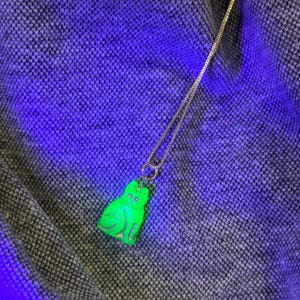 May include: A silver necklace featuring a neon green cat pendant. The cat pendant has etched details and glows brightly. The necklace chain is thin and delicate, with a small circular clasp. The background is a textured grey fabric.