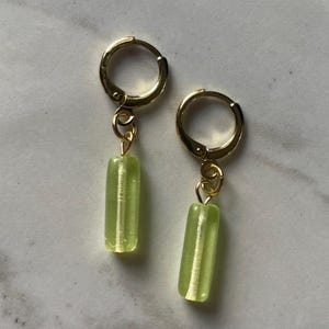 May include: A pair of gold-colored hoop earrings with a small, light green, rectangular gemstone pendant. The earrings are displayed on a light-colored surface.