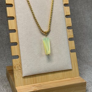 May include: A gold-colored chain necklace with a rectangular, pale green pendant. The pendant is suspended from a delicate chain and displayed on a wooden jewelry stand. The necklace is a simple, elegant piece.