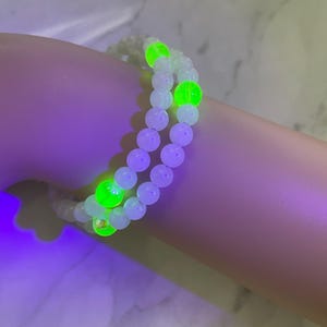 May include: A double-strand bracelet featuring white beads interspersed with glowing green beads. The bracelet is shown on a wrist, with the beads appearing to glow brightly. The bracelet is a piece of jewelry.