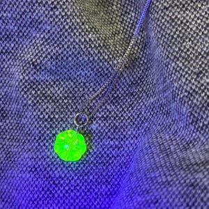 May include: A silver chain necklace featuring a neon green, flower-shaped pendant. The pendant has a textured surface and glows brightly. The necklace is displayed on a textured, dark grey and blue fabric.