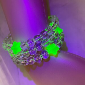 May include: A clear beaded bracelet with three rows of clear beads and bright green star-shaped accents. The bracelet is shown on a light-colored surface with a purple and green glow.