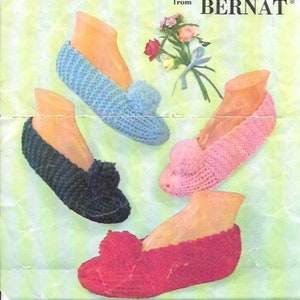 May include: Four pairs of knitted slipper mocs in blue, black, pink, and red. The mocs are made with Bernat yarn and are designed to be knit or crocheted. The image includes the text "Cuddle-Mocs to knit or crochet from BERNAT".