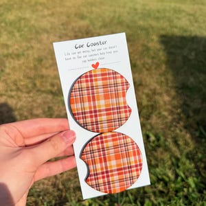 May include: Two car coasters with a plaid pattern in shades of orange, red, and yellow. The coasters are on a white card with the text "Car Coaster" and a message about keeping cup holders clean.