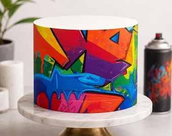 May include: A cylindrical cake featuring a vibrant graffiti-style design with bold splashes of red, orange, yellow, purple, and blue. The cake sits on a white marble cake stand with a gold-toned base, positioned near a black spray paint can.