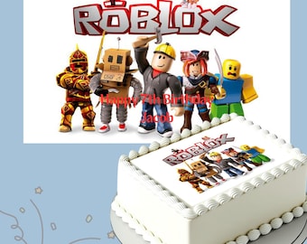 May include: A white rectangular cake decorated with a Roblox-themed image and characters. The cake is topped with white frosting and a decorative border. The image includes the Roblox logo and the text "Happy 7th Birthday Jacob."