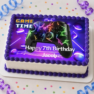 Puede incluir: Una tarta de cumpleaños rectangular con glaseado morado y base blanca. La tarta está decorada con un diseño de mando de videojuegos negro y las palabras "Game Time" y "Happy 7th Birthday Jacob". Serpentinas y confeti coloridos rodean la tarta.