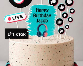 May include: A round birthday cake decorated with TikTok-themed toppers. The cake has a speckled white frosting and is adorned with colorful decorations, including a 'Happy Birthday Jacob' message, music notes, and social media icons.