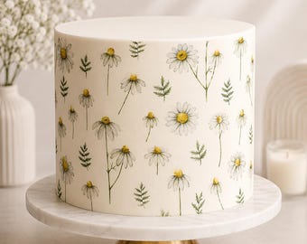 May include: A cylindrical cake with a smooth white frosting surface decorated with a repeating watercolor-style pattern of white daisy flowers with yellow centers and green stems and leaves, displayed on a white marble cake stand.