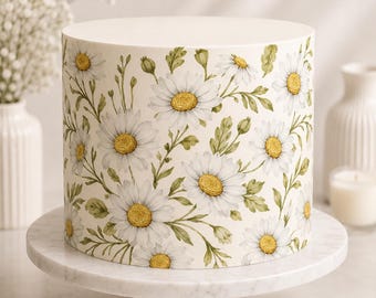 May include: A cylindrical cake with a smooth white frosting base, decorated with a repeating pattern of white daisy flowers featuring yellow centers and green stems with leaves, displayed on a white marble cake stand with a gold metal base.