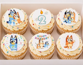 May include: Six cupcakes with white frosting and colorful sprinkles, each topped with a circular edible topper featuring Bluey characters, a large number 2 with a candle, or the text Happy Birthday in multicolored letters on a white background.