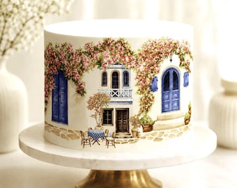 May include: A cylindrical cake featuring a wrap-around illustration of a Mediterranean-style building with blue doors, a balcony, and pink flowering vines. A small table with a blue and white checkered tablecloth and two chairs sits in front.