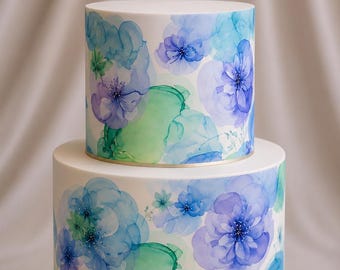 May include: A two-tier cake with a smooth white finish, decorated with a watercolor-style floral pattern in shades of blue, purple, and green. The design features layered, translucent flower petals and abstract ink-like washes of color.