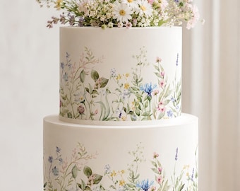 May include: A two-tier white cake decorated with a watercolor-style botanical print of wildflowers and greenery. The top of the cake is adorned with a fresh arrangement of daisies, lavender, cornflowers, and baby's breath in shades of blue, pink, and white.