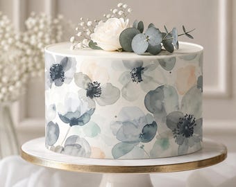 May include: A round cake with a floral design in shades of blue, gray, and peach. The cake is decorated with a white rose, sprigs of baby's breath, and eucalyptus leaves. It sits on a white cake stand with a gold rim.