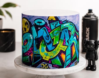 May include: A cylindrical cake featuring a vibrant graffiti-style wrap with bold blue, green, yellow, and purple spray-paint patterns. The design includes stylized lettering and abstract shapes, displayed on a white marble cake stand.