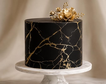 May include: A cylindrical cake with a matte black finish featuring gold veining patterns that mimic marble. The top of the cake is decorated with a cluster of metallic gold flowers and small spherical accents, resting on a white marble cake stand.