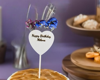 May include: A white frosted cake with caramel topping, topped with a heart-shaped cake topper. The topper has the text "Happy Birthday Milana" and is decorated with blue and purple flowers. Other desserts are in the background.