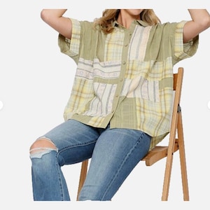 May include: A patchwork button-down shirt in shades of green and yellow, with short sleeves. The shirt features a mix of plaid and solid fabric panels. Worn with blue jeans with a distressed knee, the shirt is paired with a wooden folding chair.